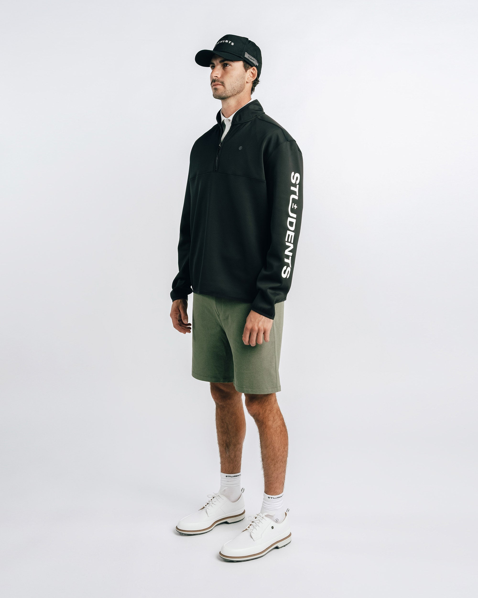 Man wearing a black sweatshirt with 'STUDENTS' text, green shorts, and white sneakers on a white background