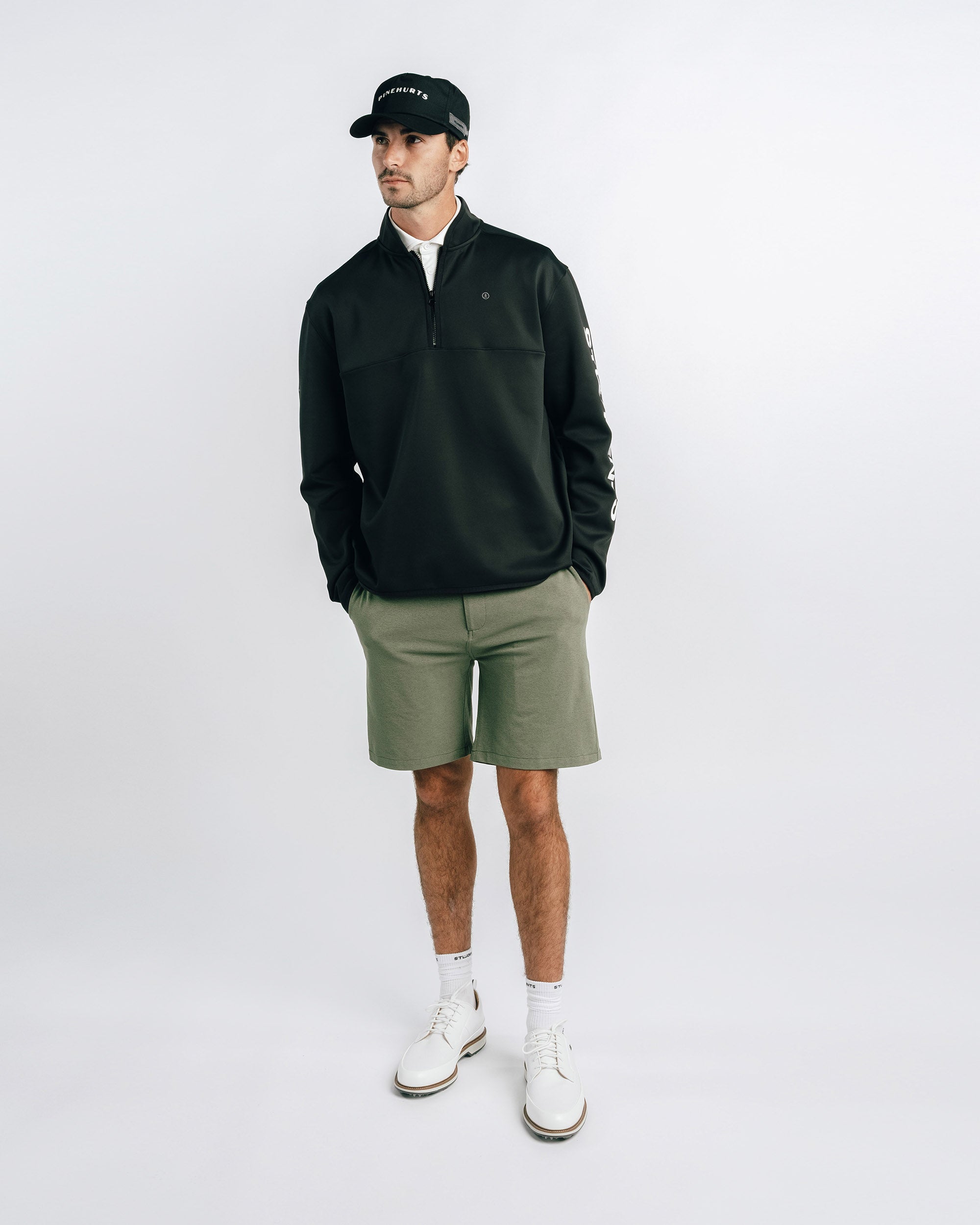 Man wearing a black jacket, green shorts, white sneakers, and a black cap on a white background
