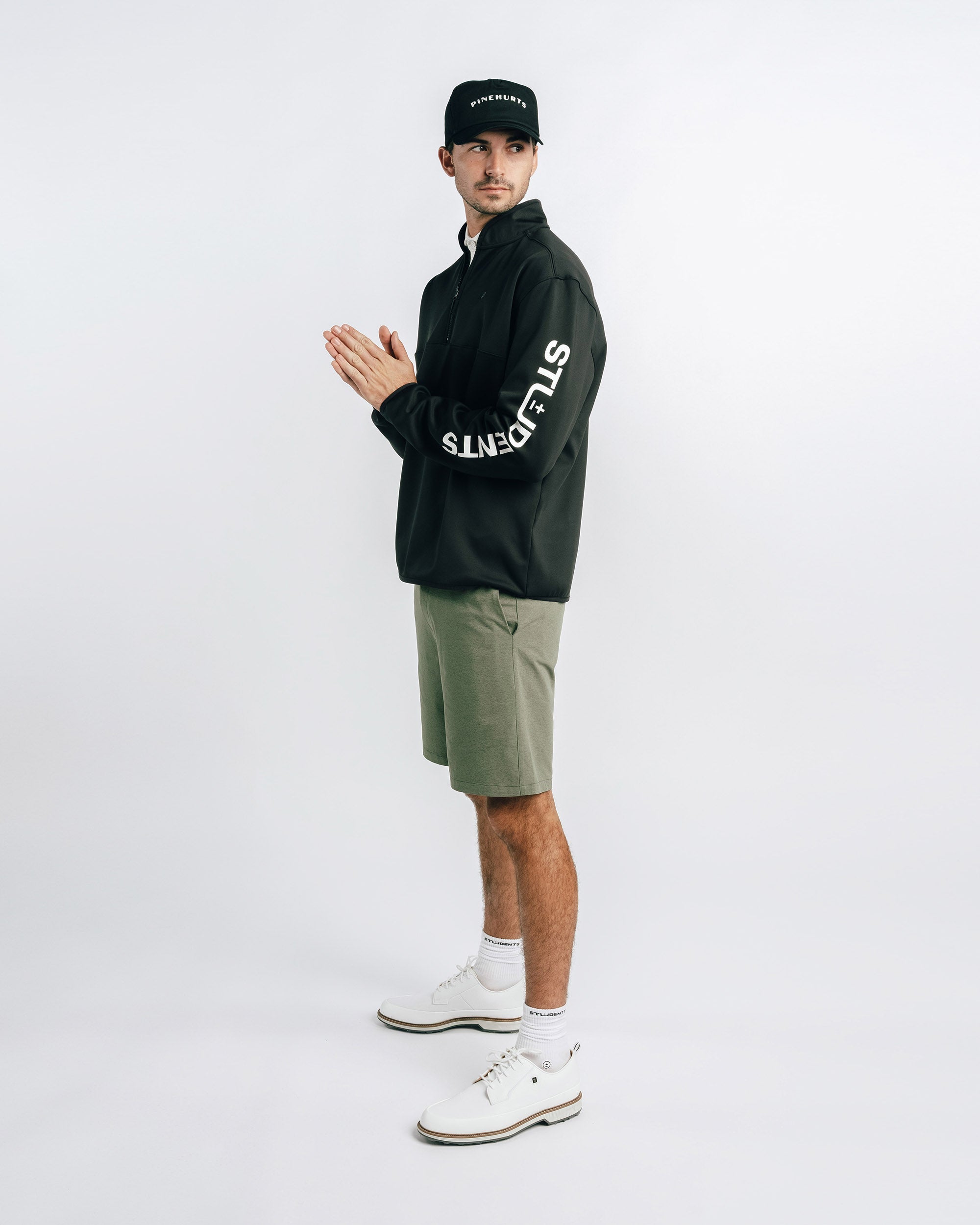 Man wearing a black jacket with white text, green shorts, and white shoes on a white background