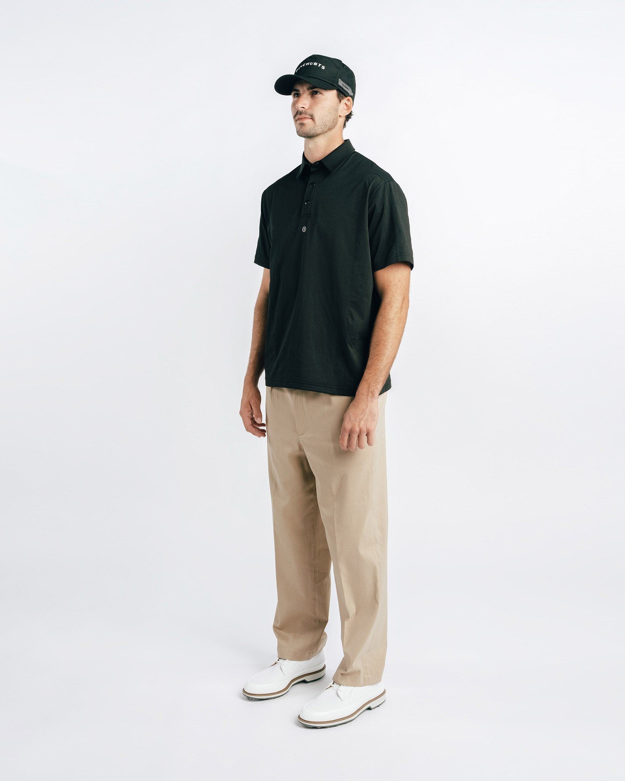 Man wearing a black polo shirt, beige pants, and a black cap on a white background