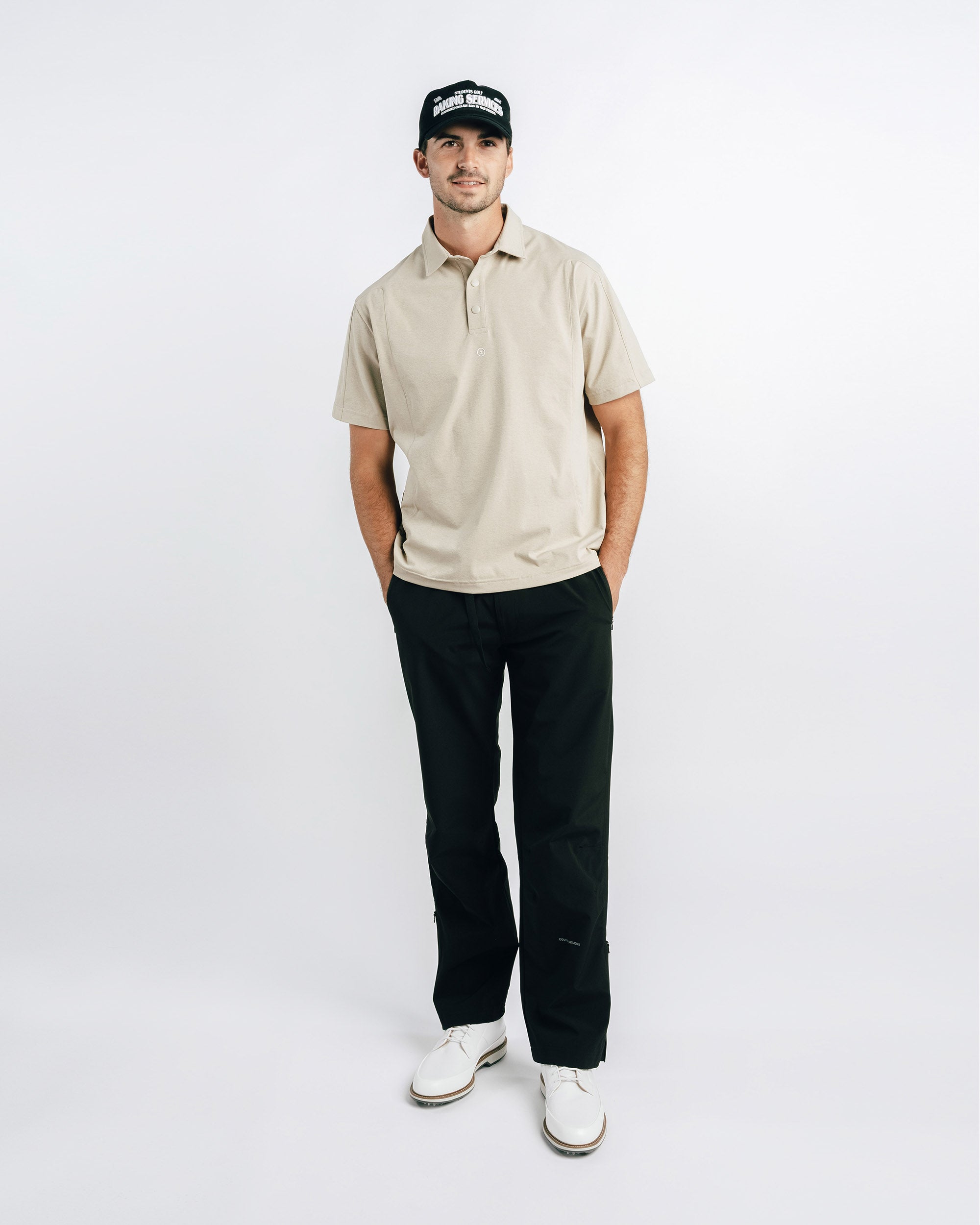 Man wearing a beige polo shirt, black pants, and a cap on a white background