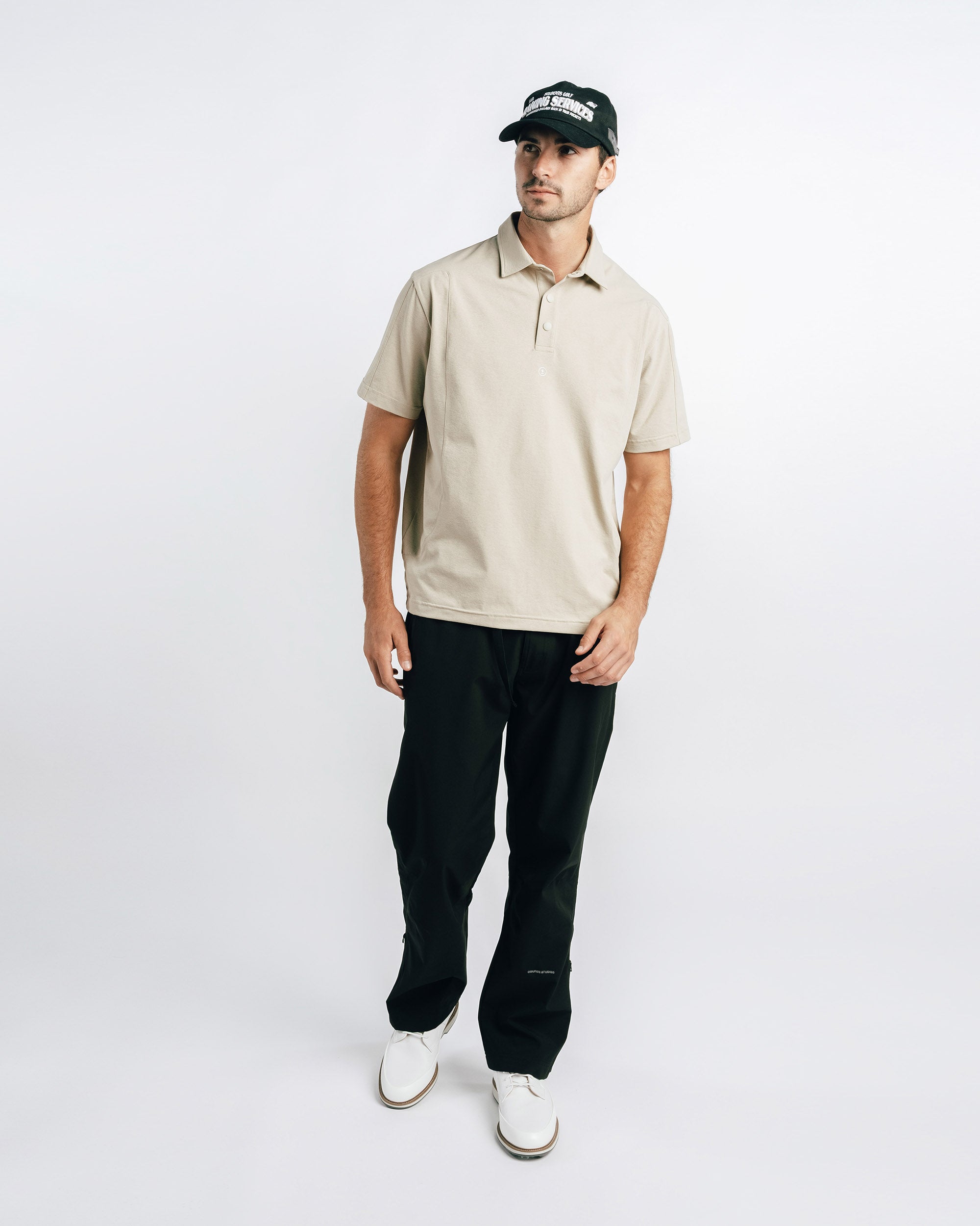 Man wearing a beige polo shirt, black pants, and a black cap on a white background