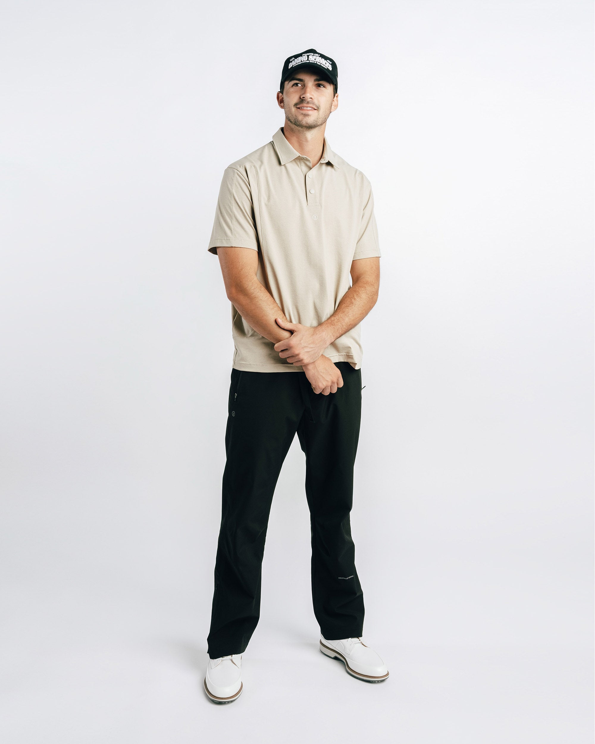 Man wearing a beige polo shirt, black pants, and a cap on a white background