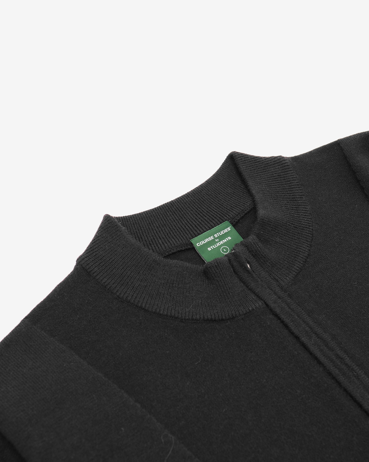 Close-up of a black ribbed knit sweater with a mock neck collar, featuring a green brand tag that reads 'GUSTAVI & LARS EST. 2016'.