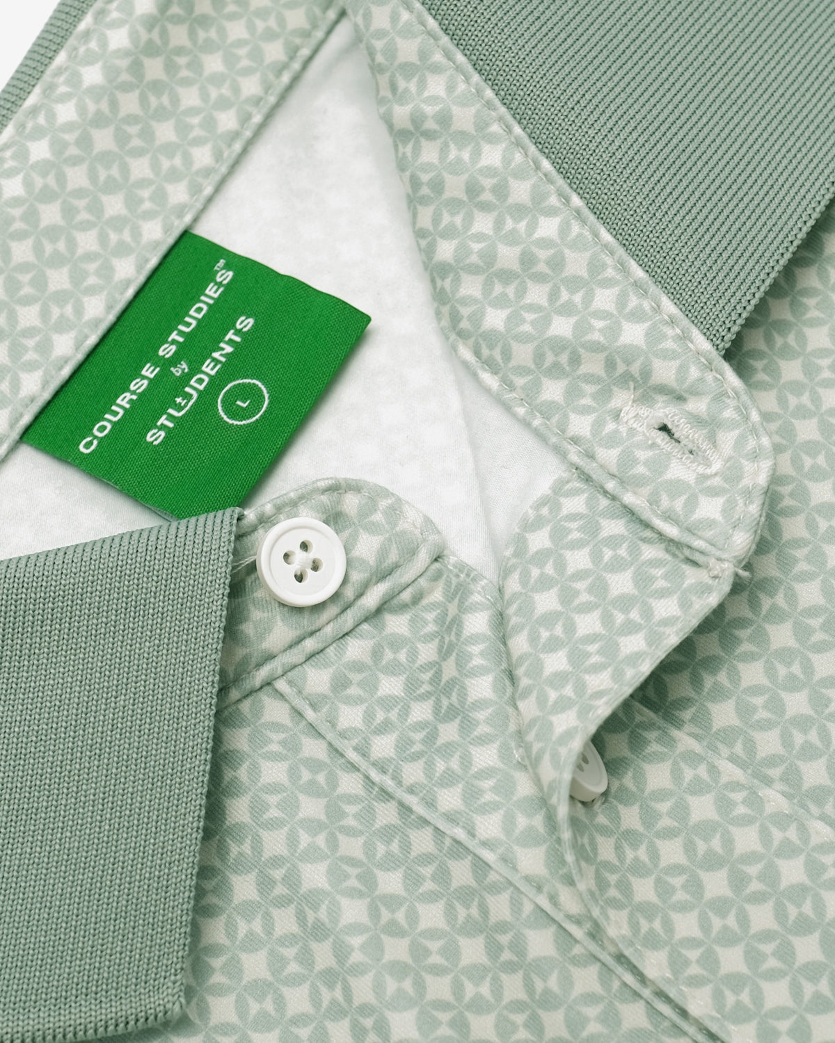 Close-up of a light sage green polo shirt with a repeating geometric pattern, featuring a solid green neck label that reads 'COVERAGE STUDENTS' and a white button.