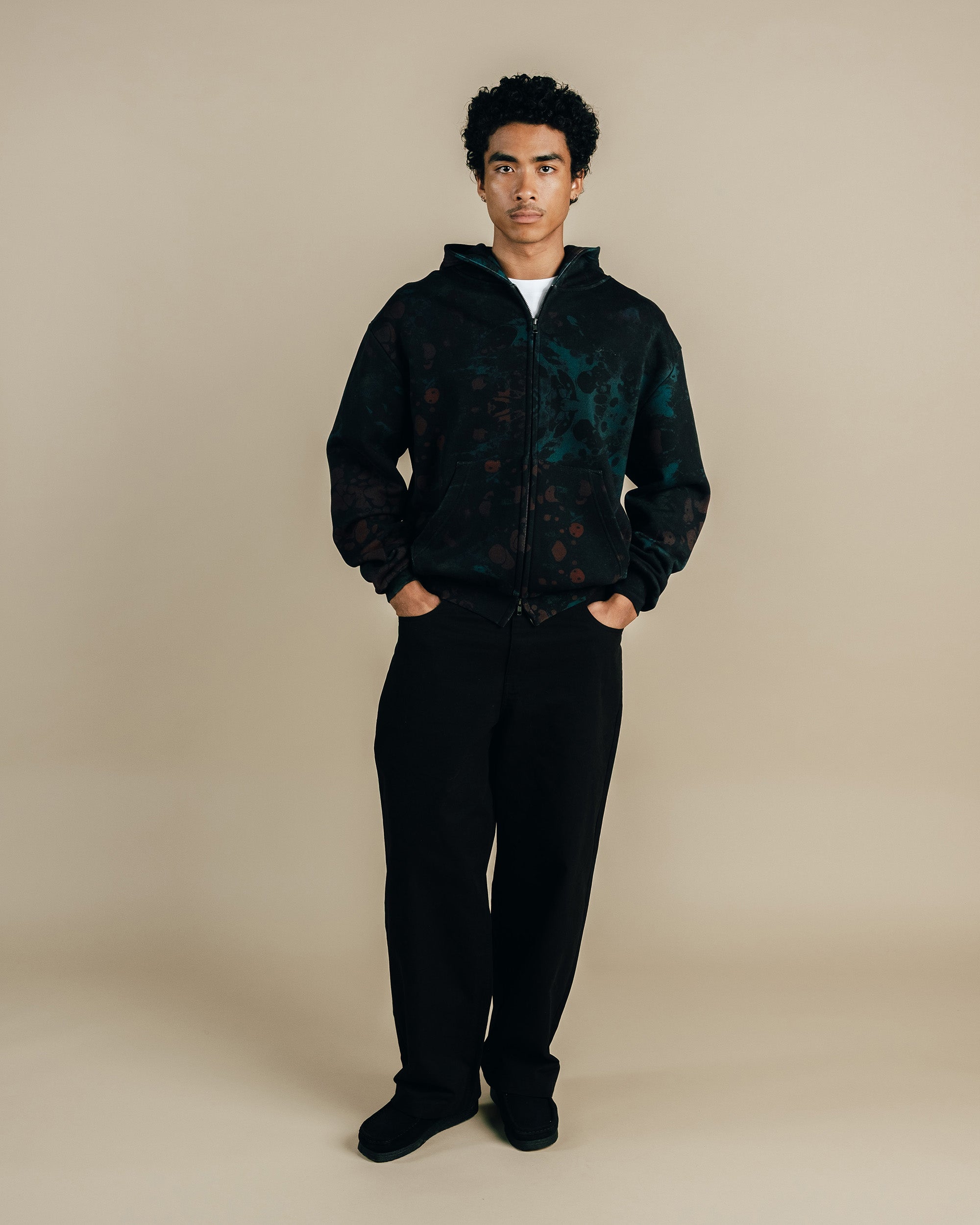 Man wearing a dark patterned jacket and black pants against a beige background