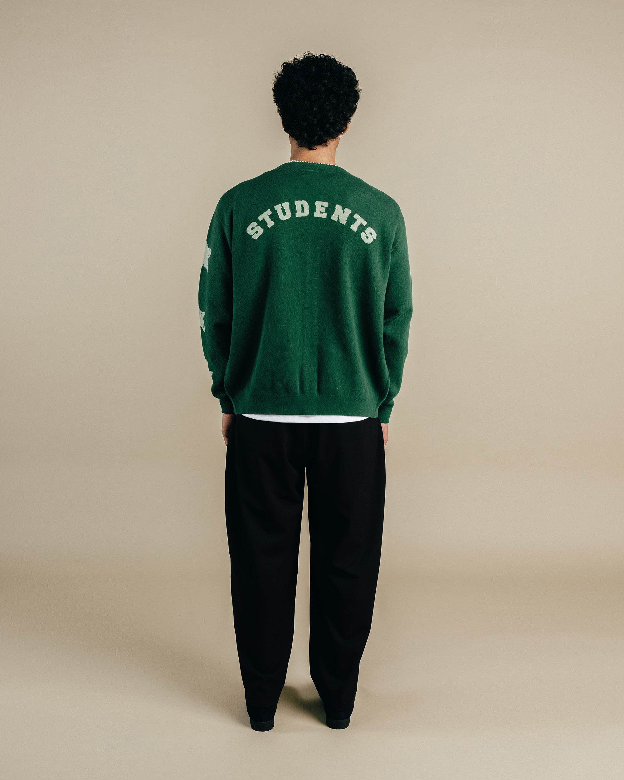 Person wearing a green sweatshirt with 'STUDENTS' text on the back, standing against a beige background.