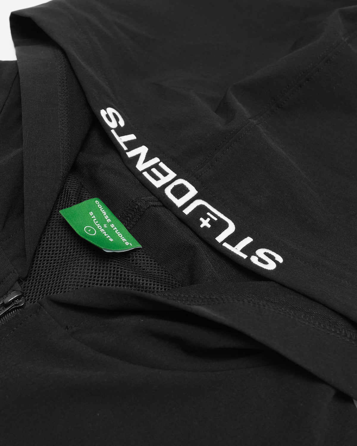 Close-up of a black zip-up hoodie or jacket with large white 'STUDENTS+' text printed along the shoulder and a green fabric brand tag visible inside the collar.