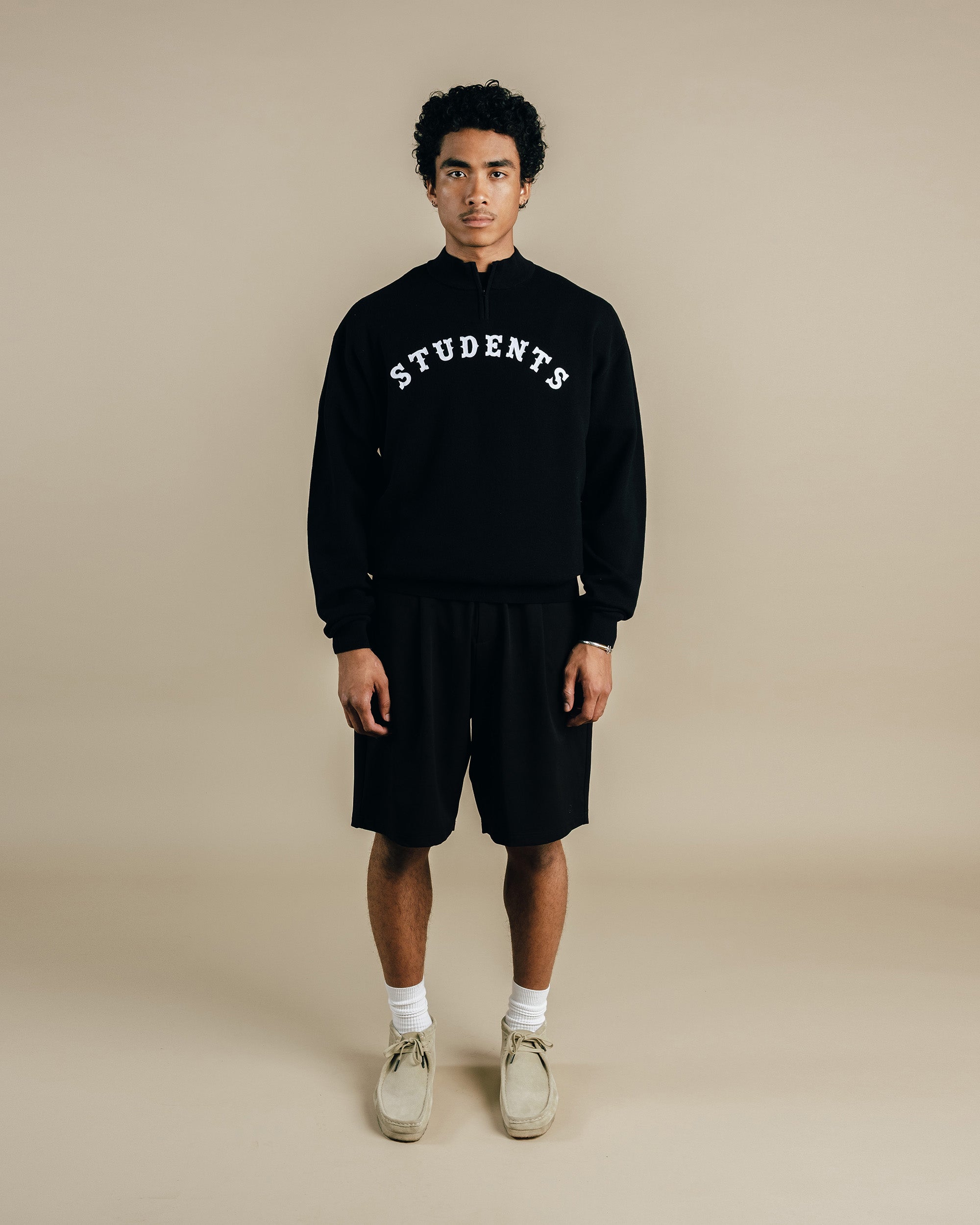 Person wearing a black sweatshirt with 'STUDENTS' text and black shorts on a beige background