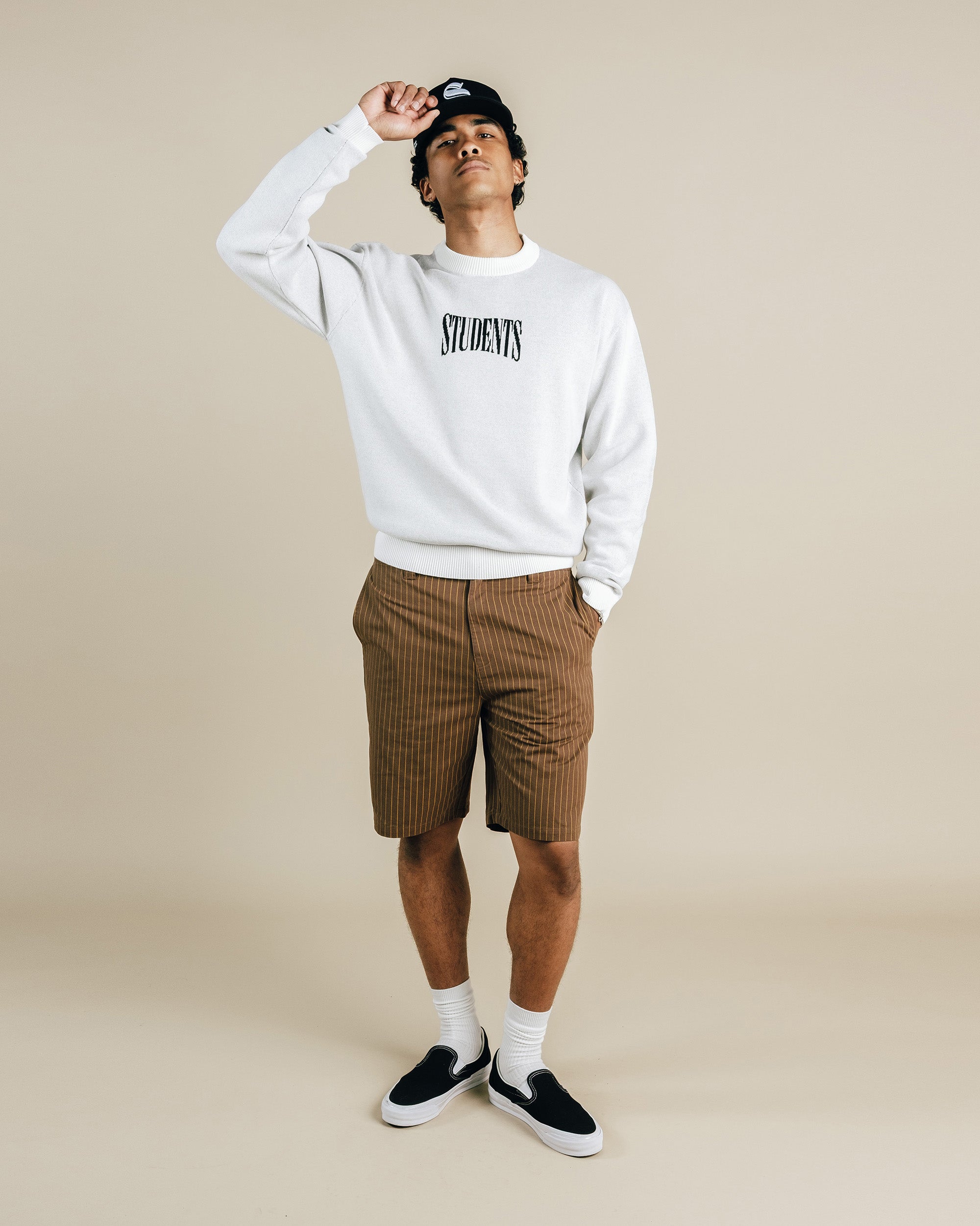 Person wearing a white sweatshirt with 'STUDENTS' text, brown checkered shorts, and black shoes on a beige background