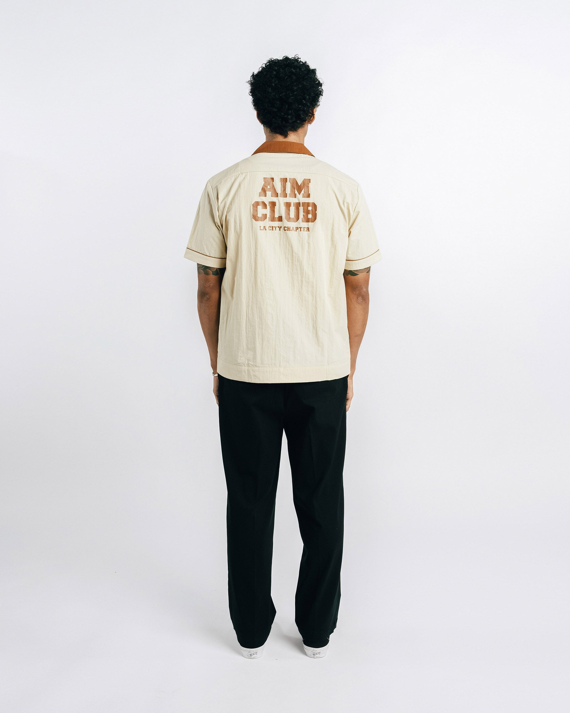 Person wearing a beige t-shirt with 'AIM CLUB' text on the back, standing against a white background