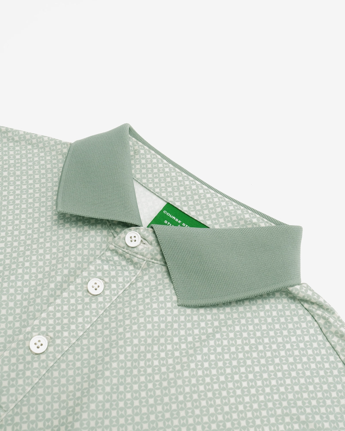 Close-up of a light sage green polo shirt with a repeating geometric pattern, featuring a solid green collar and white buttons.