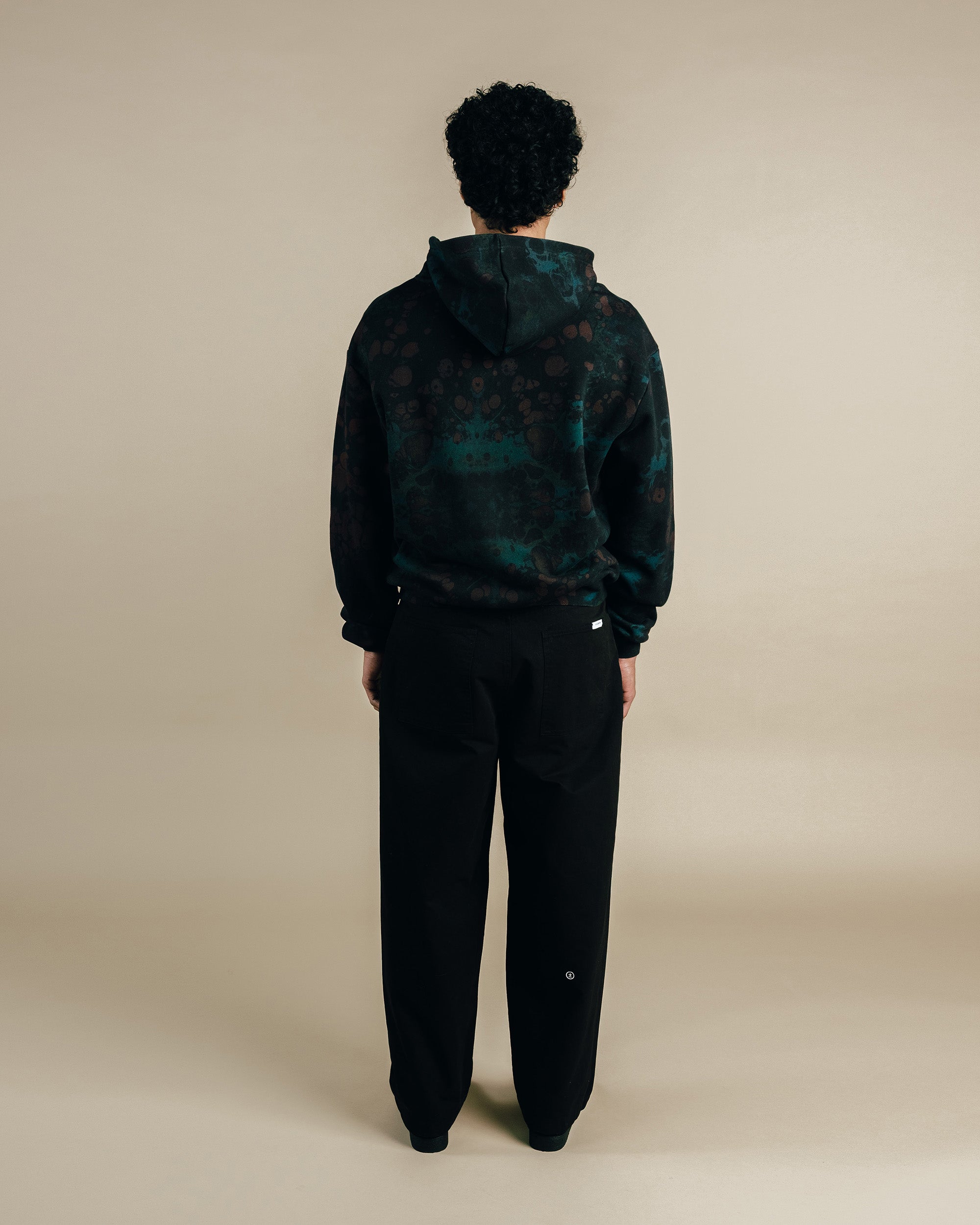 Person wearing a dark patterned hoodie and black pants on a beige background
