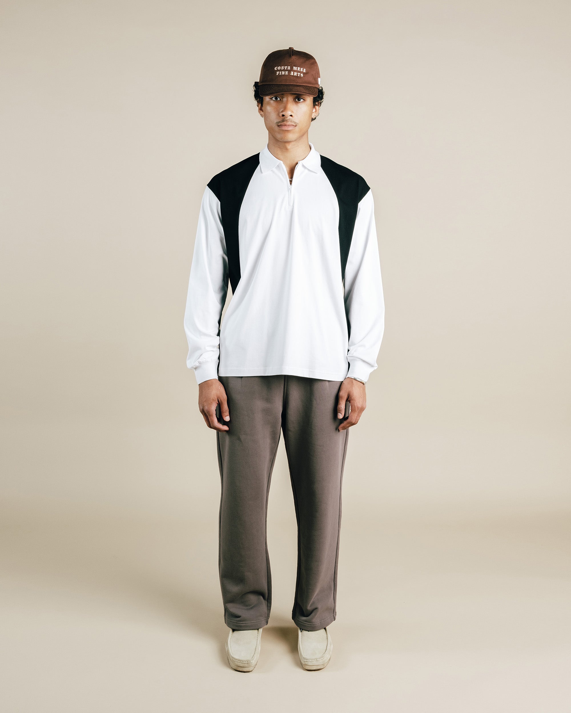 Person wearing a white shirt with black accents, brown pants, and a brown cap on a beige background