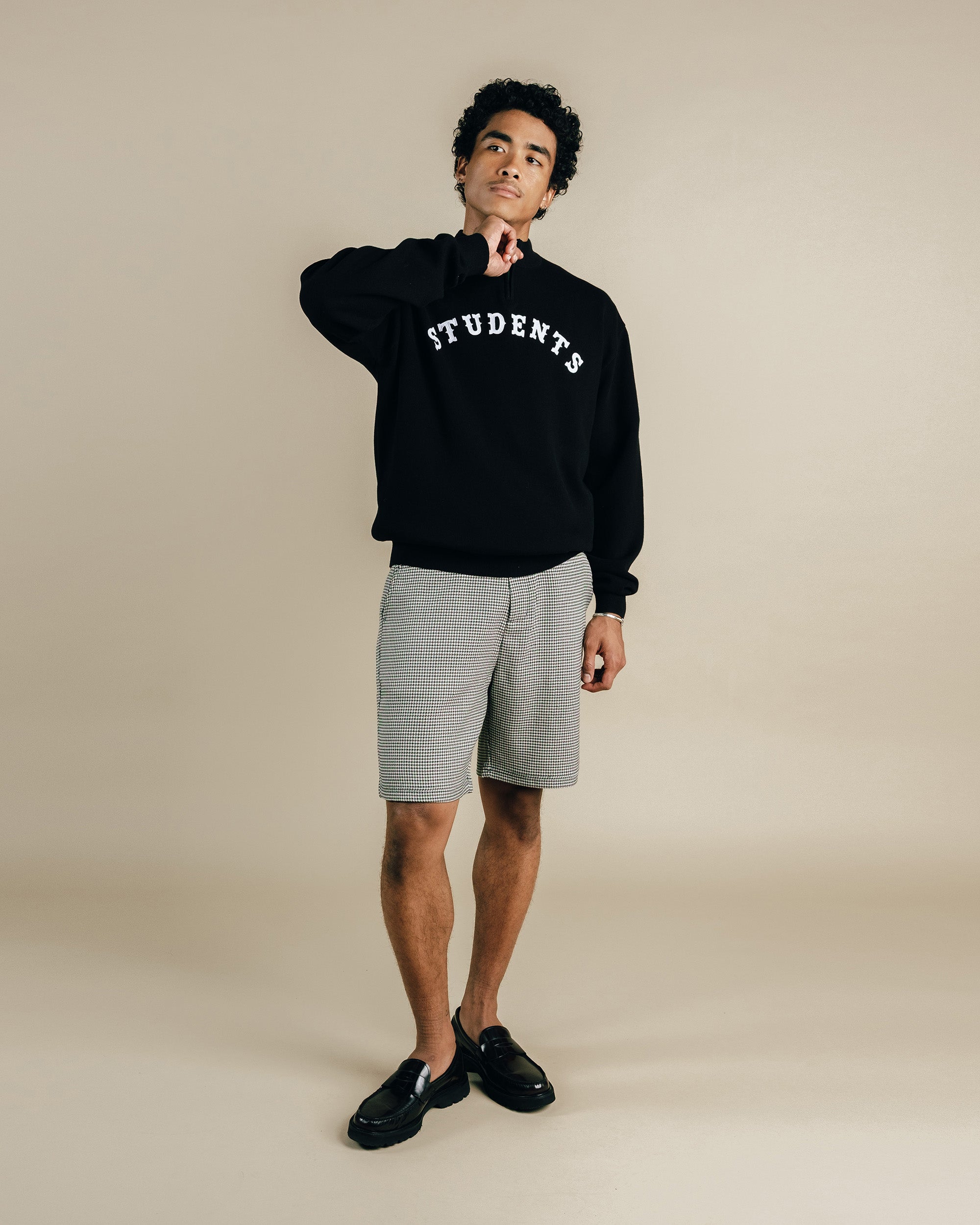 Person wearing a black sweatshirt with 'STUDENTS' text and gray shorts on a beige background