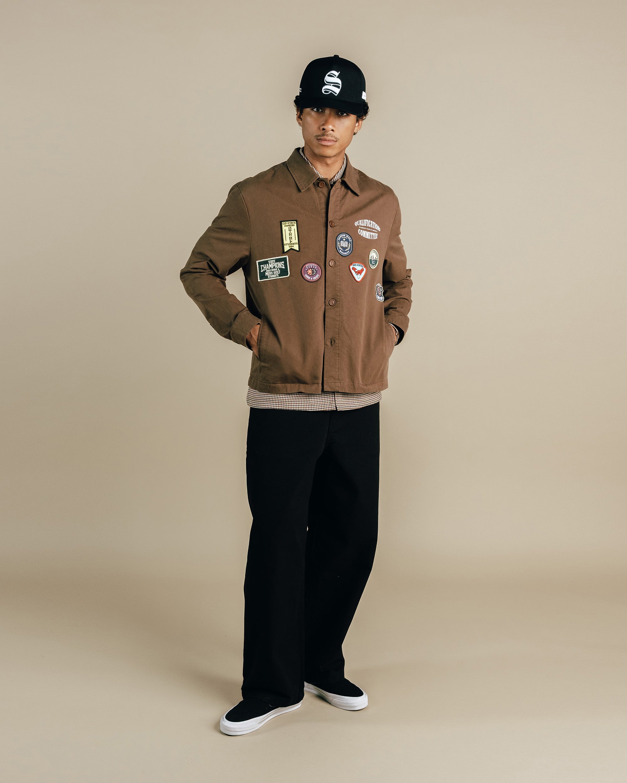 Person wearing a brown jacket with patches, black pants, and a black cap on a beige background