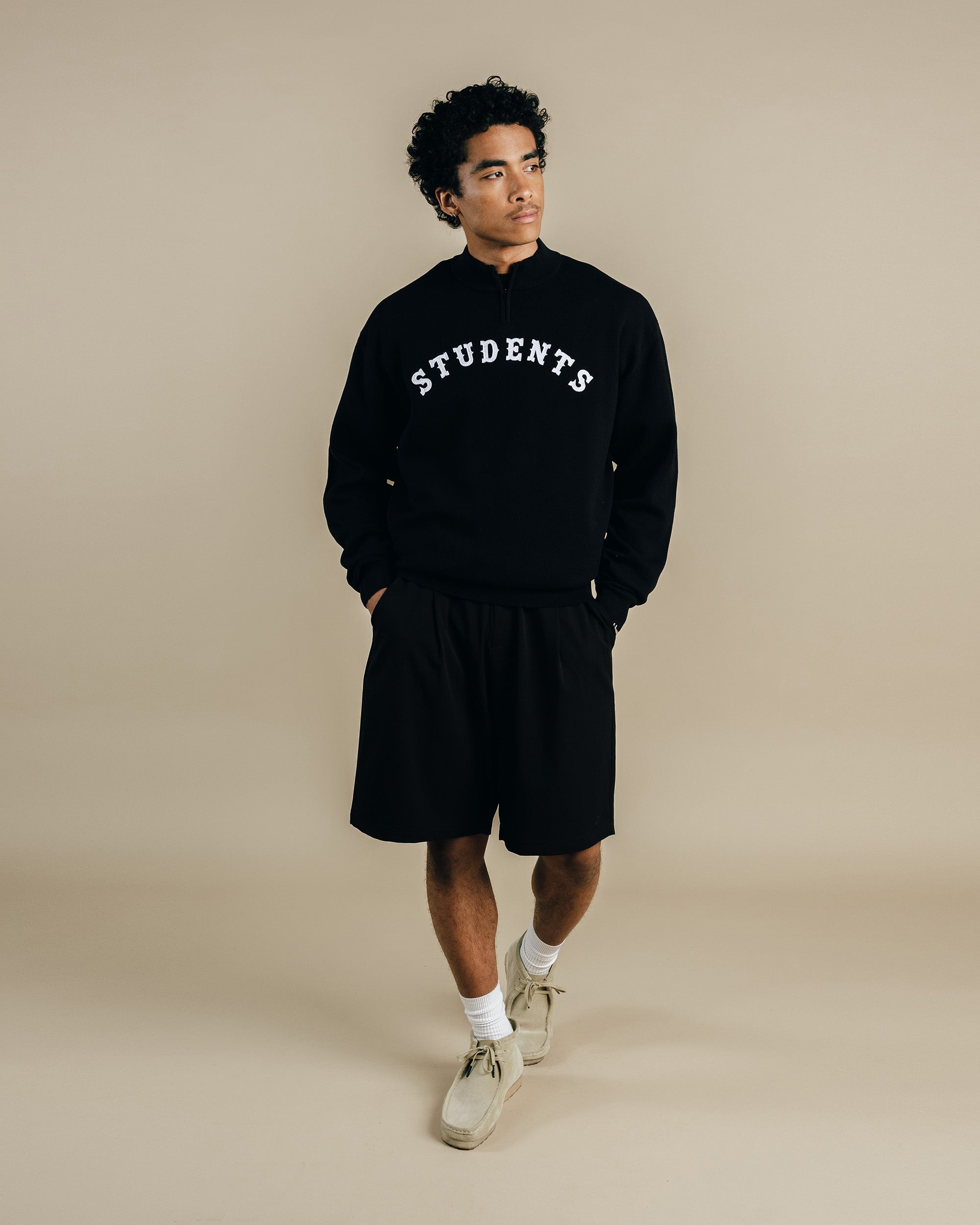 Person wearing a black 'STUDENTS' sweatshirt and shorts on a beige background
