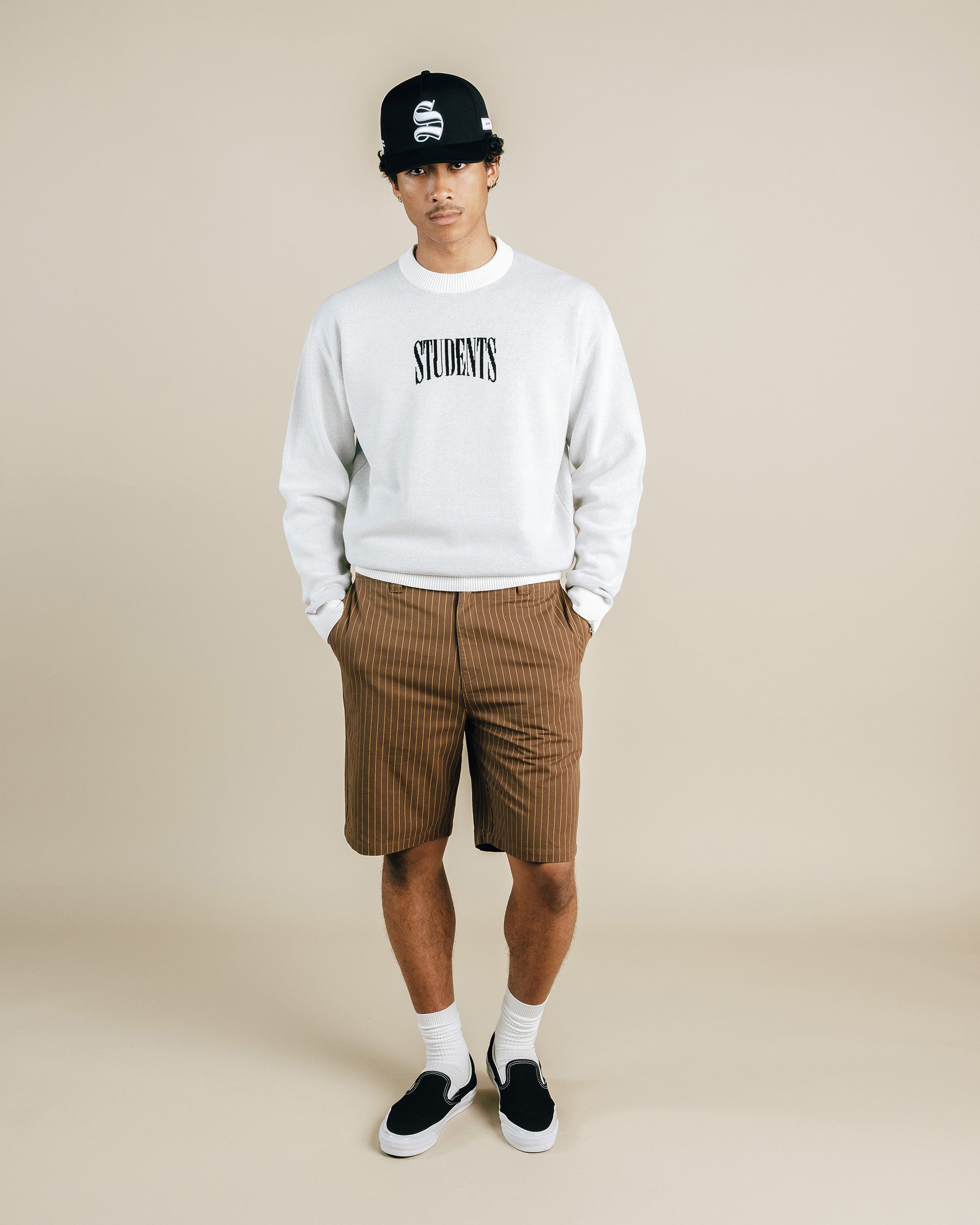 Person wearing a white sweatshirt with text, brown plaid shorts, and black sneakers on a beige background